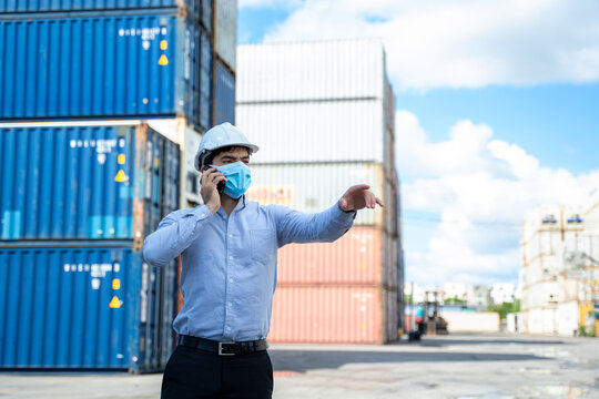 Business man wearing protective mask to Protect Against Covid-19 worker checking container box with use smart phone in shipping yard, Control loading unloading container box freight.