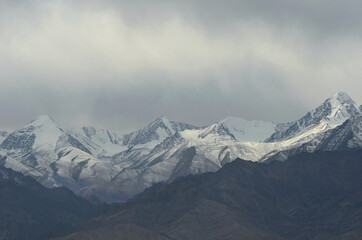 Snow covered mountain peaks rise into grey storm clouds. A shaft of sunlight falls onto one patch of snow. Rocky mountain slopes are in the foreground.