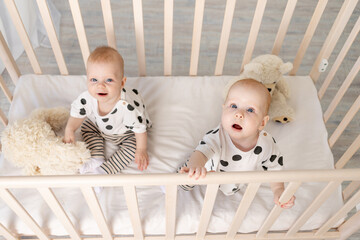 two baby twins brother and sister 8 months sit in their pajamas in the crib and look at the camera,...