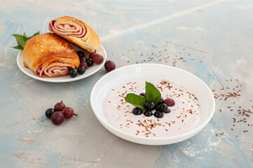 Breakfast on a blue concrete background. A plate of yoghurt decorated with currants and gooseberry berries and a fresh bun with fruit filling. Copy space.
