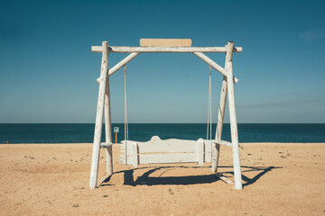 Swing on the seashore. Swing-bench on a sandy beach next to the sea. White swing on the beach. Summer vacation at the sea. Chill-out at sea.