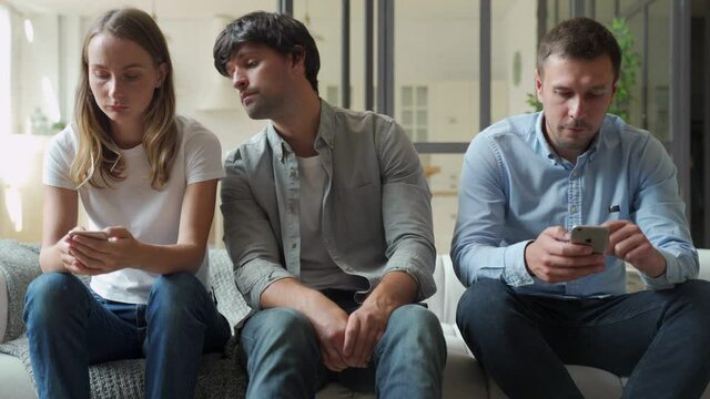Bored man sitting on couch between gadget addicted friends with smartphones