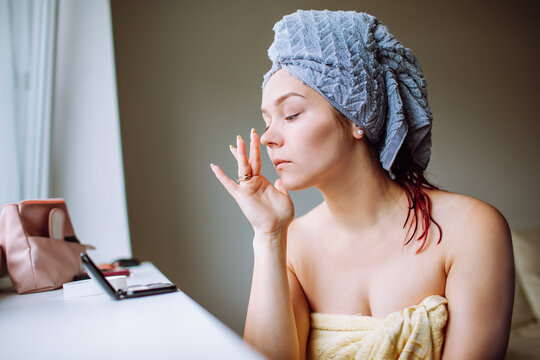 Beautiful young woman with blue towel on her head applying anti-wrinkles cream at her face while looking to the small mirror.