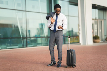 Black Entrepreneur Man Using Cellphone Standing With Luggage At Airport