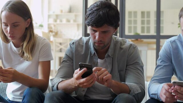 Group of three friends hanging out but ignoring each other while using their smartphones at home