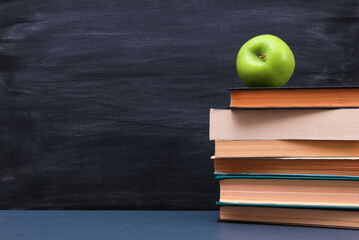 Green apple on stacked books on dark blue desk with blackboard and chalk streaks in background. Side view, copy space, close-up. Learning, education, knowledge, library, love reading concept