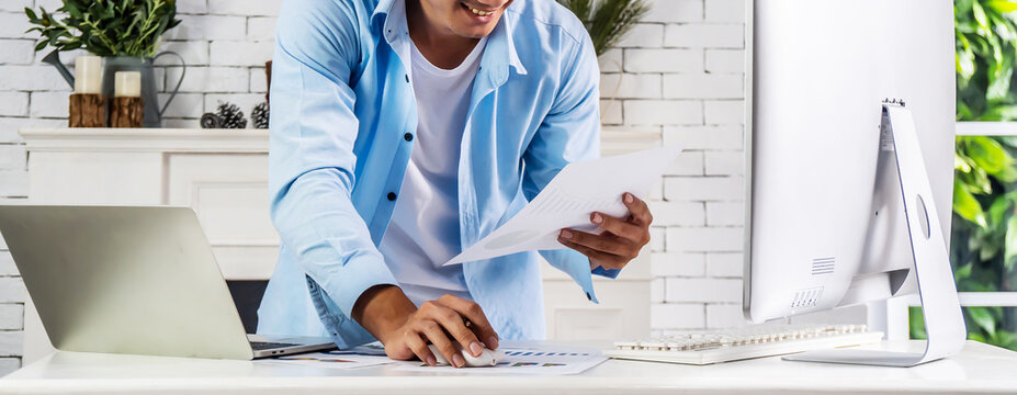 Young businessman smiling happy asian man working holding a piece of paperwork and reading while standing in front of laptop at home, work from home concept
