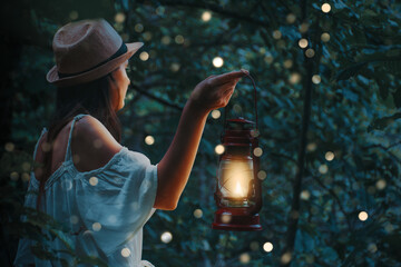 Beautiful woman in white dress holding a lantern in a forest with blurred glowings under the trees. - obrazy, fototapety, plakaty