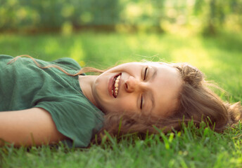 Naklejka na ściany i meble Happy laughing fun casual kid girl lying on the grass on nature summer background. Closeup positive