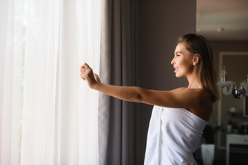 Beautiful woman wearing white towel after shower, open window cover curtains to see morning sun light, enjoy moment at luxury hotel.