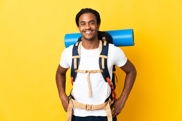 Young mountaineer man with braids with a big backpack isolated on yellow background posing with arms at hip and smiling