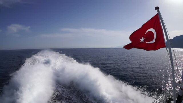 The flag of Turkey flies at the stern of the pleasure boat against the backdrop of the blue sea, mountains and the foam trail of the ship's engine.