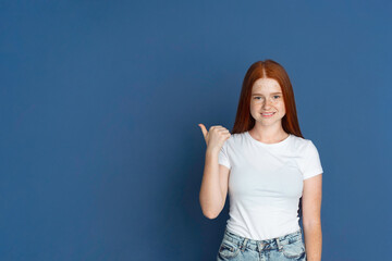 Fototapeta na wymiar Pointing at side. Caucasian young girl's portrait isolated on blue studio background. Beautiful female redhair model with cute freckles. Concept of human emotions, facial expression, sales, ad, youth