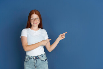 Pointing, choosing. Caucasian young girl's portrait on blue background. Beautiful female redhair model with cute freckles. Concept of human emotions, facial expression, sales, ad, youth culture.