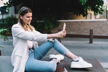 Happy millennial in earphones for listening audio using modern cellphone gadget for clicking selfie pictures during leisure time, pretty hipster girl shooting video vlog while blogging on mobile