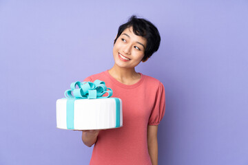 Young Vietnamese woman with short hair holding a big cake over isolated purple background looking up while smiling