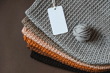 Stack of knitted material from threads of brown, orange, gray colors with an empty price tag on a brown background. Top view. Copy, empty space for text
