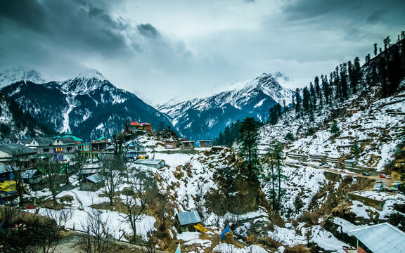 A landscape of a snow capped valley in a mountain with pine trees and houses. Blue sky with clouds above