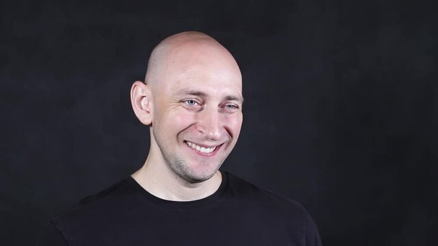 A bald caucasian man in a black t-shirt on a black background depicts winks one eye close-up. High quality FullHD footage