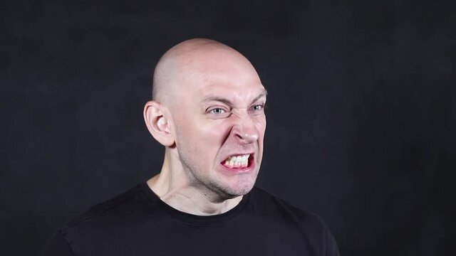 A bald Caucasian in a black T-shirt on a black background depicts a grimace of anger close-up. High quality FullHD footage
