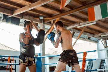 Training in the gym in the ring with a muay thai trainer. Tattooed caucasian man and Asian trainer....