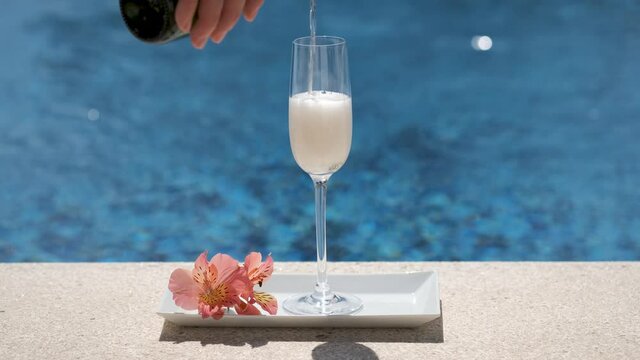Close up of pouring glass with champagne or prosecco on swimming pool or sea background. Waiter pours cold champagne into glass. Sparkling white wine on tray with flowers. Luxury alcohol drink. 4K