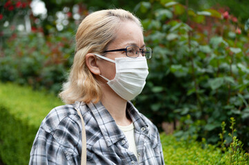 Side view portrait of middle aged mixed race blonde woman with eyeglasses wearing white surgical mask. Protection against coronavirus (COVID-19) and other infectious diseases. Outdoors. Copy space.