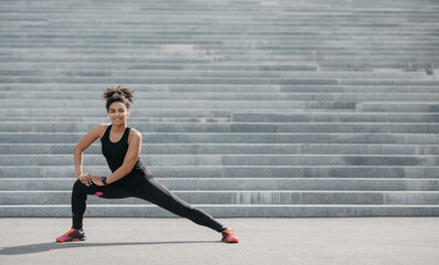 Fototapeta na wymiar Fit exercises after jogging. Smiling african american girl in sportswear doing stretching for legs on gray stairs background