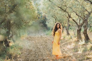 Woman in yellow summer linen dress in olive tree garden. Portrait of beautiful curly brunette girl...