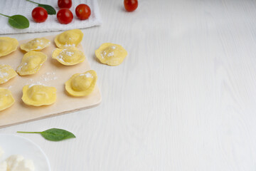 Raw italian ravioli pasta filled with ricotta cheese surrounded by spinach and tomatoes on towel on white wooden background. Image with copy space, horizontal orientation