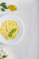 Top view of italian ravioli pasta filled with cheese in a plate decorated by ricotta and basil leaves surrounded by spinach and olive oil gravy boat on white wooden background. Image with copy space