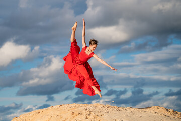 young ballerina in a bright red long dress soars in a jump above the ground, like a bird, against a...