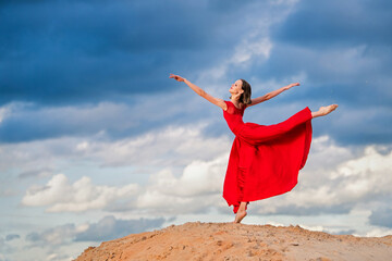young ballerina in a bright red long dress stands in an arabesque, like a bird, against a...