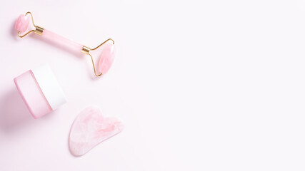 Pink quartz facial roller, gua sha and moisturizing cream on pastel pink. Tools for skin care routines, top view, copy space