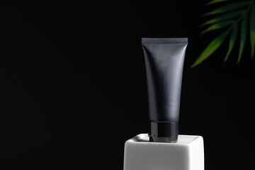 Black unbranded cosmetic tube with cream, mask or lotion on white ceramic podium on black background with copy space for your design. Face care concept.