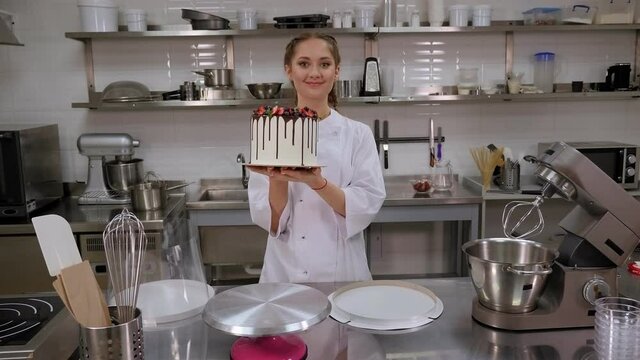 A female pastry chef holds a cake with chocolate streaks, strawberries and blueberries in a professional pastry kitchen. Portrait of a pastry chef with a cake in his hands.