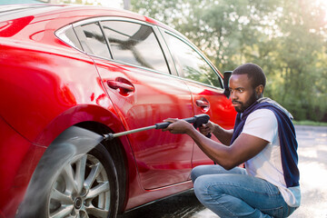 Young African man in casual wear washing wheels of his luxury car at self car wash station...