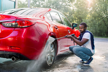 Young African man in casual wear washing wheels of his luxury car at self car wash station outdoors, using high pressure water jet. Car wash outdoors concept
