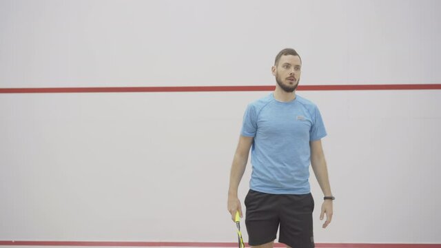Portrait of serious handsome personal trainer controlling workout of unrecognizable sportsman in gym. Brunette young Caucasian man serving ball and talking as blurred guy playing squash at front.