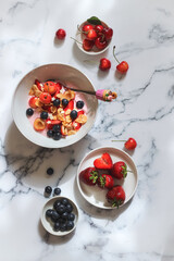A bowl of yogurt with strawberries, blueberries, cherries and flakes on a marble table