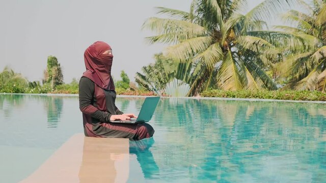 Muslim freelancer working on laptop while sitting in swimming pool outdoors.