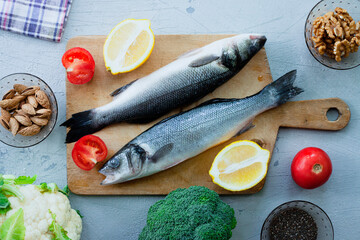 Fresh raw fish sea bass on cutting board, raw vegetables, spices and nuts. Stone background. Healthy food, lifestyle, diet and weight control concept. Space for a text.
