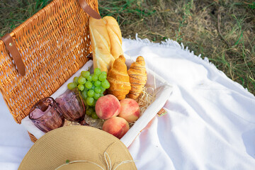Picnic basket on grass with food and drink on blanket. Picnic lunch outdoor in a field on sunny day with baguettes, croissants, peaches, a bouquet of wildflowers and grapes. French style picnic