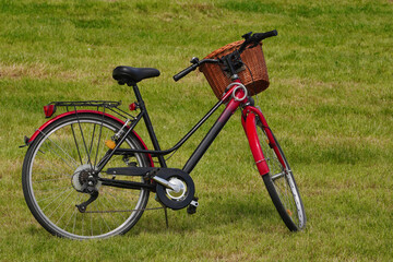 Fototapeta na wymiar A red-and-black city Bicycle with a basket stands in a meadow, on the newly mown grass in the Park. Walking and driving in the fresh air in Sunny weather, an eco-friendly form of transport
