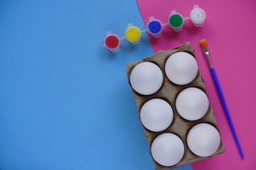 Tray or box of six eggs on a blue and pink background, multi-colored paints and brushes. Set for easter DIY children's creativity. Minimal easter concept. Symbol Happy Easter Flat lay