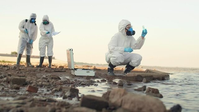 Wide shot of group of three scientist exploring polluted seashore. Female scientist taking water sample while her two colleagues having talk on background
