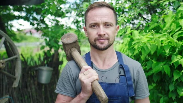 Worker in the garden. Young adult man stands in the summer garden with hammer and looks at the camera