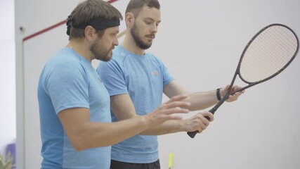 Professional trainer explaining how to hold squash racket to mid-adult Caucasian man. Portrait of confident young coach training brunette bearded guy in gym. People playing squash sport game.