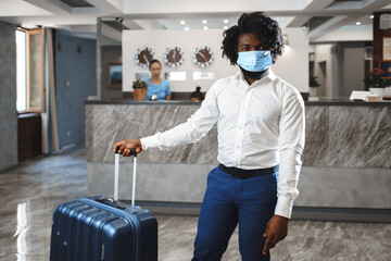 African man hotel guest with suitcase wearing protective mask to protect from coronavirus