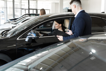 Side view of young beautiful woman sitting inside car and holding hand on steering wheel. She smiling and talking with manager of car dealership. Car agent representing inside of automobile.